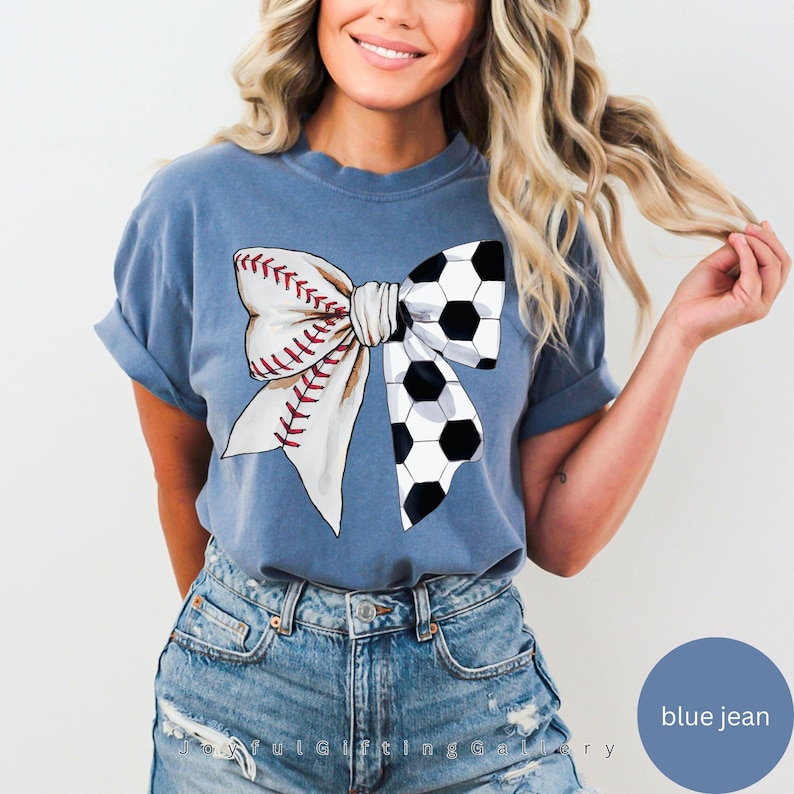 May include: A blue jean t-shirt with a white bow design. The bow is made up of a baseball and a soccer ball.