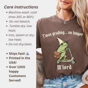 May include: Brown t-shirt with a green frog riding a scooter, wearing a wizard hat and carrying a satchel. Text on the shirt reads "T'was grading... no longer" and "M'lord". Care instructions are listed on the left.