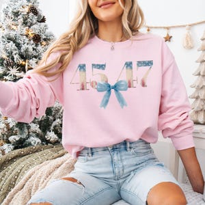May include: A pink sweatshirt with the number 4547 printed on it in a distressed American flag design. A blue bow is centered below the number.