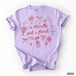 May include: A light purple t-shirt with the text "I'm literally just a florist" in red. The design includes red floral illustrations, bows, and hearts. The word "ORCHID" is printed in the bottom right corner.