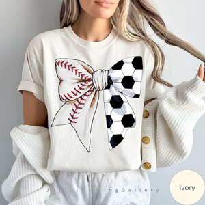 May include: A white t-shirt with a graphic of a baseball and soccer ball bow. The bow is white with red stitching and black and white soccer ball patterns. The shirt is ivory.