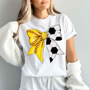 May include: White t-shirt with a yellow and black bow made from a softball and soccer ball.
