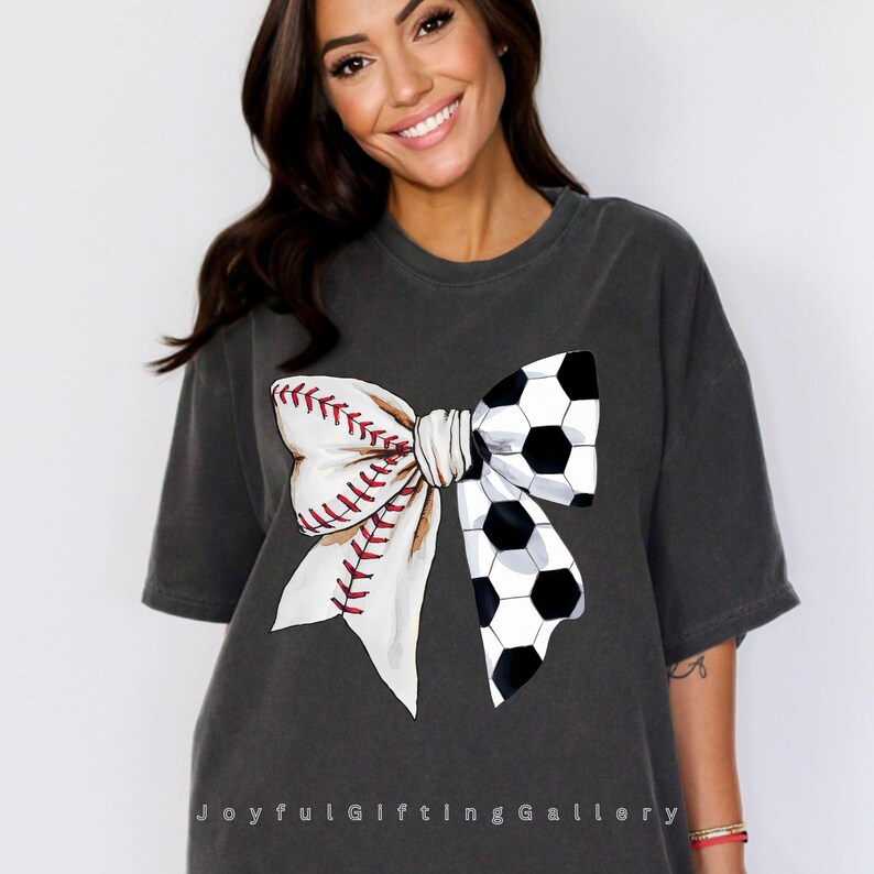May include: A gray t-shirt with a white bow made from a baseball and a soccer ball. The bow is centered on the shirt.