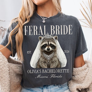 May include: Charcoal grey t-shirt featuring a raccoon in a bridal veil and crown, with the words "FERAL BRIDE". Below, it reads "OLIVIA'S BACHELORETTE Miami, Florida" and the year "2026".