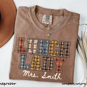 May include: A brown short-sleeve t-shirt with a crew neckline and a Comfort Colors label. The graphic features the text LUNCH LADY in a multi-coloured plaid pattern, with the name Mrs. Smith printed in white cursive script underneath the text.