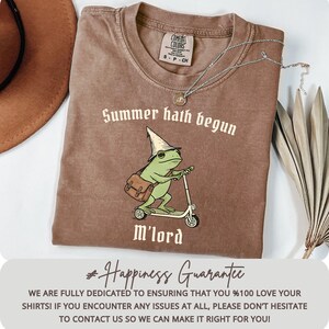 May include: Brown t-shirt with the text "Summer hath begun" and a cartoon frog riding a scooter. The frog wears a wizard hat and carries a satchel. A silver necklace with a pendant is on the shirt.