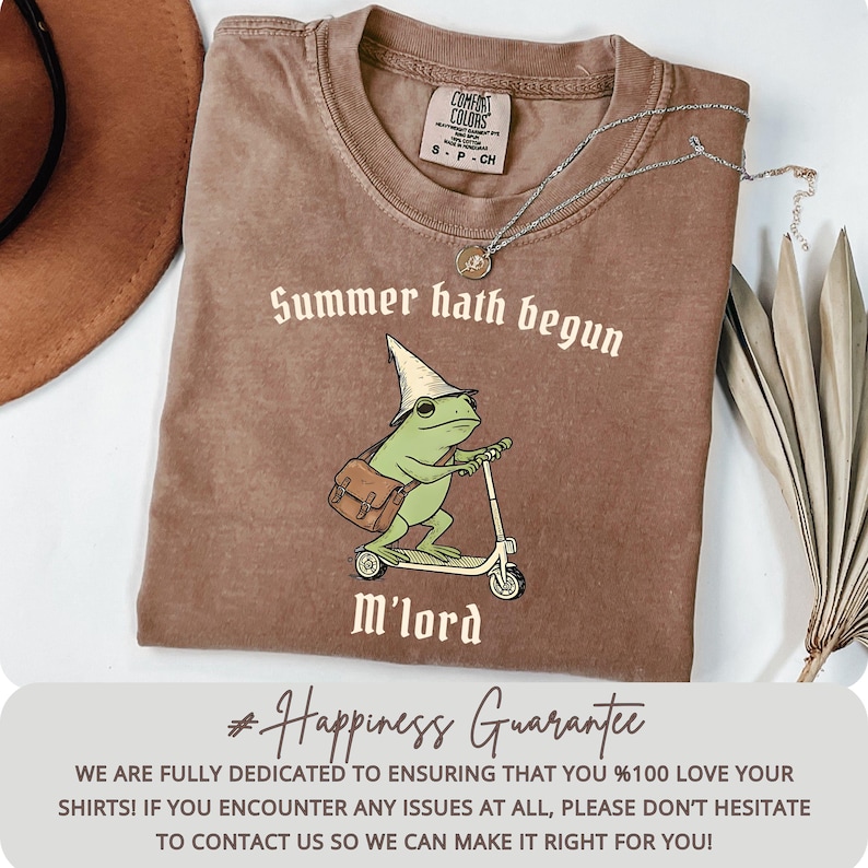 May include: Brown t-shirt with the text "Summer hath begun" and a cartoon frog riding a scooter. The frog wears a wizard hat and carries a satchel. A silver necklace with a pendant is on the shirt.