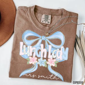 Puede incluir: Camiseta color espresso con un dise&ntilde;o de lazo azul claro y las palabras "Lunch Lady" en una fuente pastel. Debajo del texto hay flores rosas y azules. El nombre "mrs. smith" est&aacute; escrito debajo de las flores.