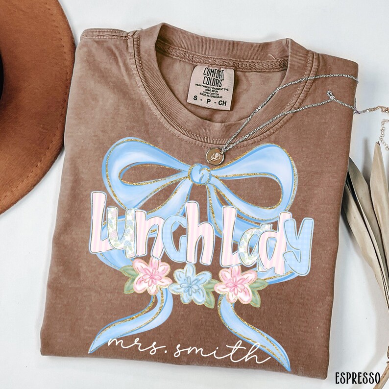 Puede incluir: Camiseta color espresso con un dise&ntilde;o de lazo azul claro y las palabras "Lunch Lady" en una fuente pastel. Debajo del texto hay flores rosas y azules. El nombre "mrs. smith" est&aacute; escrito debajo de las flores.