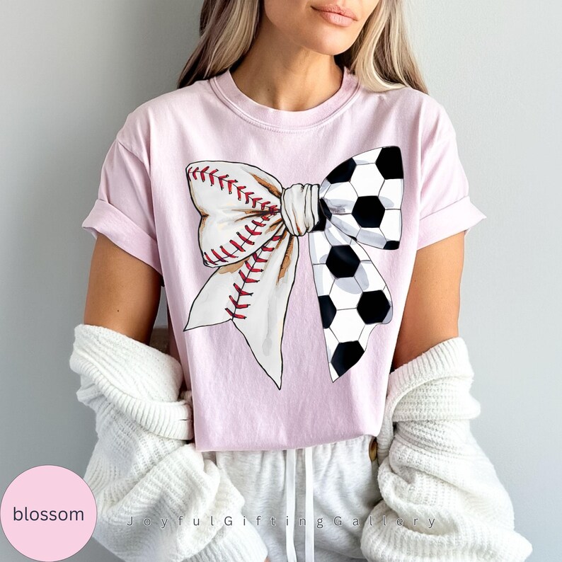 May include: A pink t-shirt with a white bow design. The bow is made of a baseball and a soccer ball.