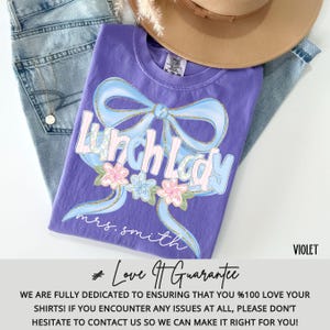 Puede incluir: Una camiseta violeta con la frase "Lunch Lady" en una fuente caprichosa, adornada con un lazo azul claro y detalles florales. El nombre "mrs. smith" est&aacute; impreso debajo. La camiseta se muestra con un sombrero y unos vaqueros.