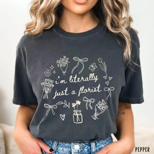 May include: Dark gray t-shirt with the text "I'm literally just a florist" surrounded by floral illustrations, including bouquets, bows, and hearts. The shirt is a comfortable, casual style.