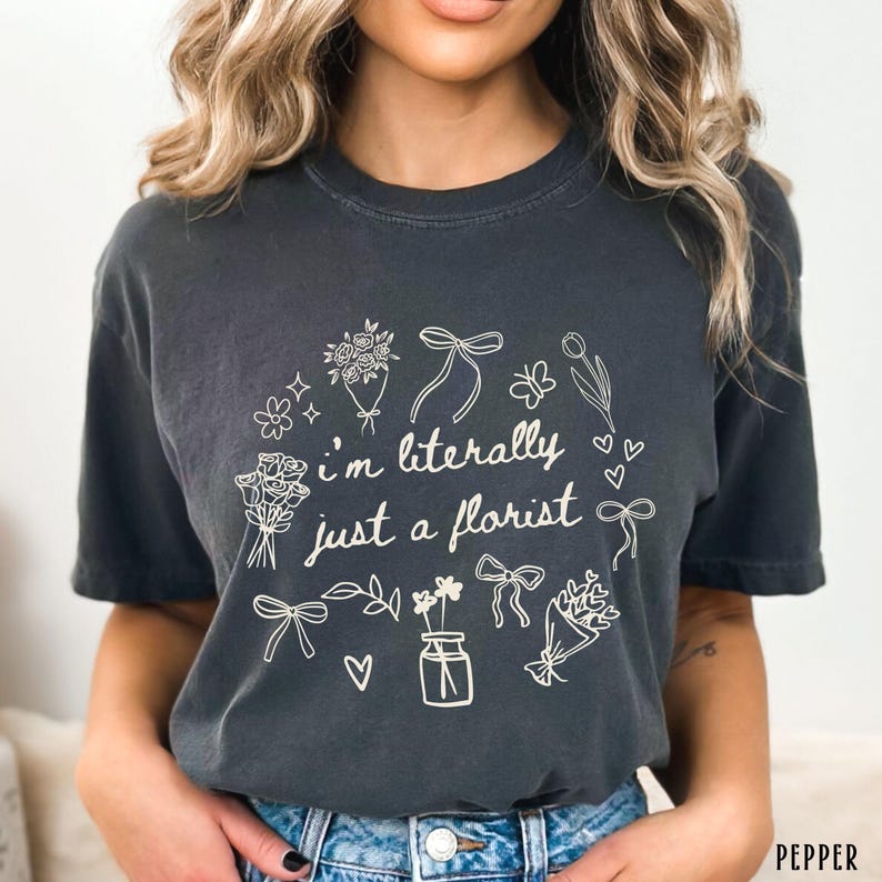 May include: Dark gray t-shirt with the text "I'm literally just a florist" surrounded by floral illustrations, including bouquets, bows, and hearts. The shirt is a comfortable, casual style.