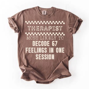 May include: Brown t-shirt with the text "THERAPIST MOST LIKELY TO DECODE 67 FEELINGS IN ONE SESSION" in white. The text is arranged in a stacked format with a checkered pattern above the word "THERAPIST".