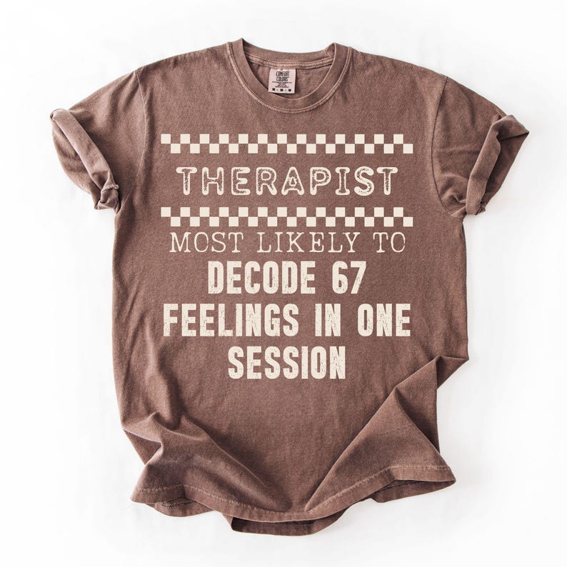 May include: Brown t-shirt with the text "THERAPIST MOST LIKELY TO DECODE 67 FEELINGS IN ONE SESSION" in white. The text is arranged in a stacked format with a checkered pattern above the word "THERAPIST".
