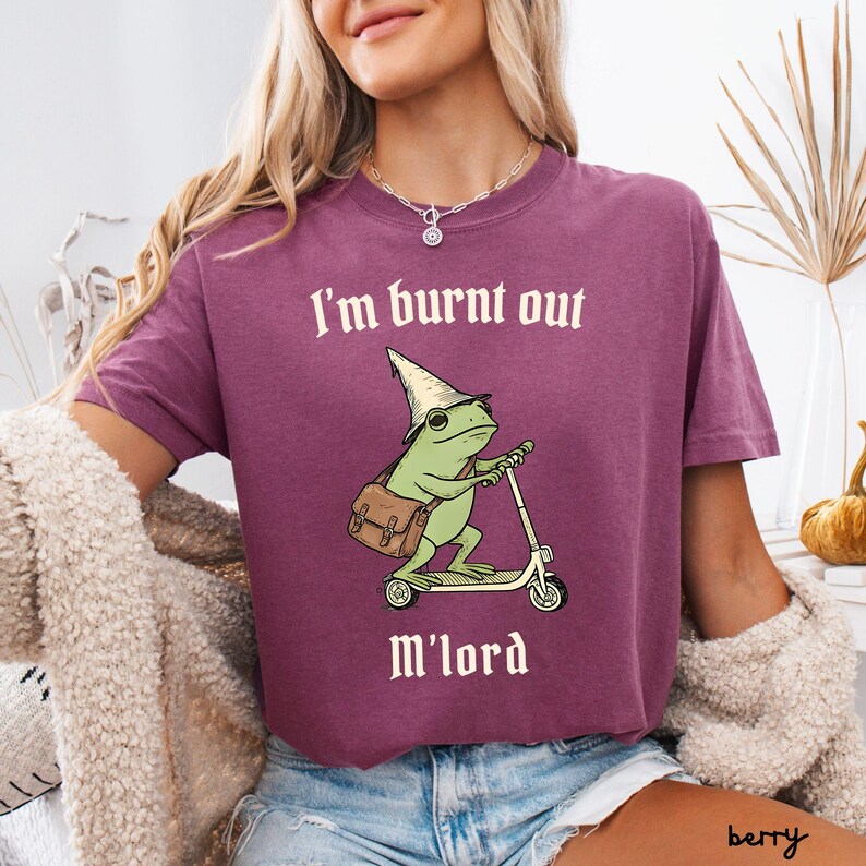 May include: Berry-colored t-shirt with the text "I'm burnt out" above a cartoon frog riding a scooter and the words "M'lord" below. The frog wears a wizard hat and carries a satchel. The shirt is a solid color.