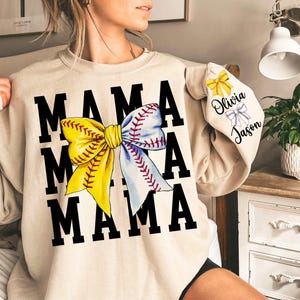 May include: A beige sweatshirt with the text "MAMA" repeated three times and a large yellow and white baseball bow in the centre. The sleeves have a smaller yellow and white baseball bow with the text "Olivia Jason" on the right sleeve.