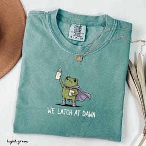 May include: A light green t-shirt featuring a graphic of a green frog wearing a purple cape, holding a baby bottle and a clipboard. The text "WE LATCH AT DAWN" is printed below the frog. The shirt has a Comfort Colors brand label at the neck.