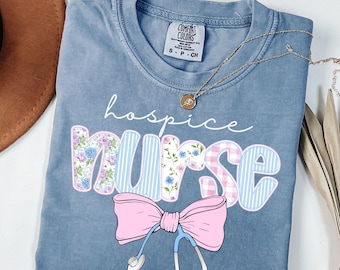 May include: A blue short-sleeve t-shirt with a Comfort Colors label. The graphic features the text hospice nurse in white script and block letters with floral, striped, and plaid patterns. A pink bow and a blue stethoscope are below the text.