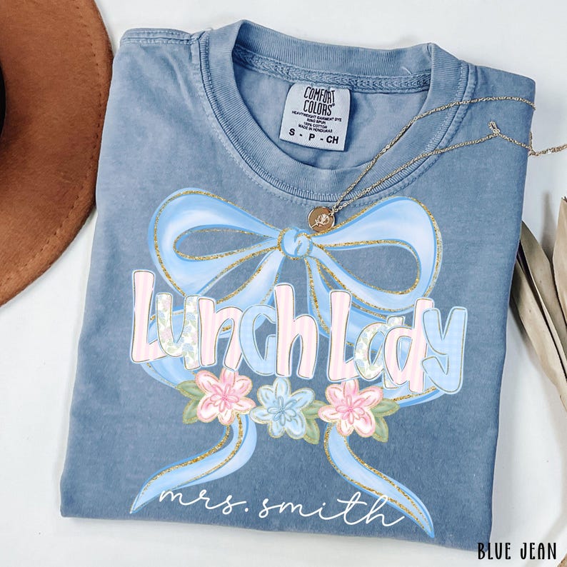 Puede incluir: Camiseta Comfort Colors azul vaquero con un dise&ntilde;o de lazo azul claro y las palabras "Lunch Lady" en rosa y azul. La camiseta tambi&eacute;n presenta el nombre "mrs. smith" en escritura blanca y un arreglo floral.