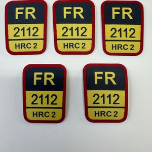 Puede incluir: Cinco parches rectangulares con esquinas redondeadas. Cada parche tiene un fondo negro con las letras "FR" en amarillo en la parte superior, seguido de "2112" y "HRC 2" en amarillo. Los parches tienen un borde rojo.