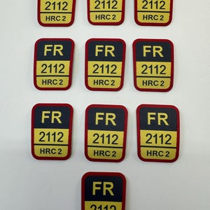 Puede incluir: Parches rectangulares con borde negro y centro amarillo. Los parches tienen las letras "FR", el número "2112" y el texto "HRC 2" impresos. Los parches están dispuestos sobre una superficie blanca.