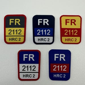 Puede incluir: Cinco parches rectangulares con esquinas redondeadas. Cada parche presenta las letras "FR", el número "2112" y "HRC 2". Los parches tienen diferentes combinaciones de colores, incluyendo rojo, amarillo, azul y negro.