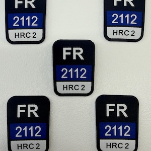 Puede incluir: Cinco parches rectangulares negros con esquinas redondeadas. Cada parche tiene las letras "FR", un rectángulo azul con "2112" en blanco y un rectángulo blanco con "HRC 2" en negro.