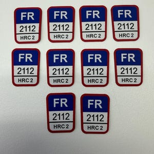 Puede incluir: Un conjunto de diez parches rectangulares. Cada parche tiene un fondo azul con las letras "FR" en blanco, seguido de "2112" y "HRC 2" en blanco. Los parches tienen un borde rojo.