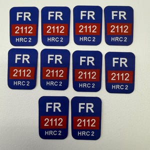 Puede incluir: Diez pegatinas rectangulares azules. Cada pegatina muestra las letras blancas "FR" en la parte superior, un rectángulo rojo con el número "2112" y el texto "HRC 2" en la parte inferior, todo en blanco.