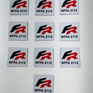 May include: Nine white square patches with a black and red "FR" logo and the text "NFPA 2112" printed below. One additional patch is shown with the same logo and text, but it is smaller than the others.