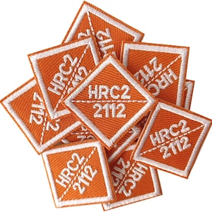 Puede incluir: Un montón de parches de tela naranja con costuras blancas. Cada parche tiene forma de diamante con el texto "HRC2 2112" en blanco.