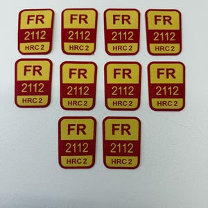 Puede incluir: Diez pegatinas rectangulares con fondo amarillo y bordes rojos. Cada pegatina tiene las letras "FR", el número "2112" y el texto "HRC 2" en negro. Las pegatinas están dispuestas sobre una superficie blanca.