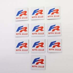 Op de afbeelding: Een verzameling van tien witte vierkante stoffen labels. Elk label heeft een blauwe en rode afbeelding en de tekst "NFPA 2112". De labels zijn gerangschikt op een witte achtergrond.