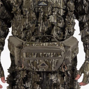 K&ouml;nnte beinhalten: Tarn-Jagdbekleidung, bestehend aus einer Jacke, Handschuhen und einer Bauchtasche. Die Jacke und die Handschuhe weisen ein blattartiges Design in Gr&uuml;n-, Braun- und Graut&ouml;nen auf. Die Bauchtasche hat mehrere Taschen und einen braunen Rei&szlig;verschluss.