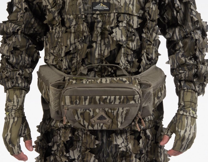 K&ouml;nnte beinhalten: Tarn-Jagdbekleidung, bestehend aus einer Jacke, Handschuhen und einer Bauchtasche. Die Jacke und die Handschuhe weisen ein blattartiges Design in Gr&uuml;n-, Braun- und Graut&ouml;nen auf. Die Bauchtasche hat mehrere Taschen und einen braunen Rei&szlig;verschluss.