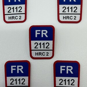 Puede incluir: Cinco parches rectangulares con fondo azul y borde rojo. Cada parche tiene las letras "FR", el número "2112" y "HRC 2". Los parches están dispuestos sobre una superficie blanca.