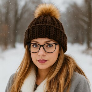 May include: A woman wearing a dark brown knit beanie with a large, fluffy, light brown pom-pom. She is also wearing black-framed glasses and a gray coat with a white fur collar. The background is a snowy, blurred outdoor scene.