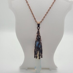 Puede incluir: Un collar con colgante de color cobre con un cristal largo, puntiagudo y opalino. El colgante tiene un diseño calado adornado con un acento azul claro. El collar se exhibe en un maniquí blanco.
