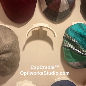 May include: A white plastic wall-mounted cap display rack with a curved support arm. The text CapCradle™ OptiworksStudio.com is printed below the central empty rack. Several flat caps in various colors and patterns are mounted on the wall.