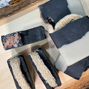 May include: Four black and white soap bars with a loofah embedded in each bar. The bars are on a wooden surface.