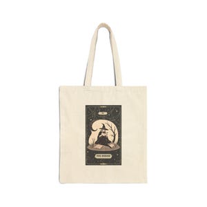 May include: A beige tote bag featuring a black and white illustration of a witch sitting on a rock with a book in her lap. The illustration is surrounded by a black frame with gold stars and the text "The Dreamer" at the bottom.