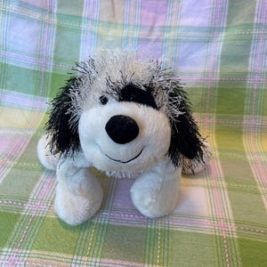 Peut inclure: Une peluche en forme de chien avec un corps blanc, une fourrure hirsute noire et blanche et un nez noir. Le chien a une expression souriante et est allongé sur une surface à carreaux.