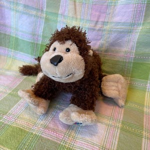 May include: A soft toy monkey with brown fur and a beige face, hands, and feet. The monkey has a black nose and is positioned on a green and white checked surface.