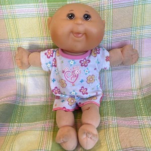 Peut inclure: Une poupée Cabbage Patch Kid vintage avec une tête chauve, des yeux bruns et une expression souriante. La poupée porte une tenue bleu clair avec un motif floral et le texte "It's a Girl!". La poupée est posée sur une couverture à carreaux.