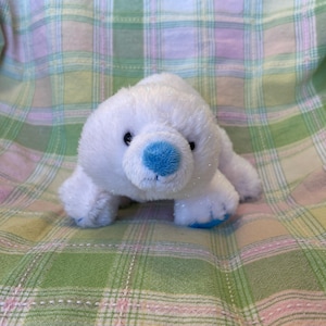 May include: A white plush polar bear toy with a blue nose and paws. The soft toy is lying on a plaid fabric background with green, pink, and white colours.