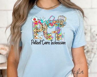 May include: Light blue t-shirt with a colorful graphic of the letters "PCT" with medical symbols and the text "Patient Care Technician".
