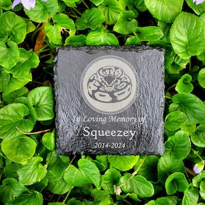May include: A square, black slate memorial stone with the text "In Loving Memory of Squeezy 2014-2024". The stone features a circular design with a snake illustration.