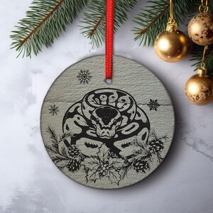 May include: Round Christmas ornament with a black and white snake design. The ornament has a red ribbon for hanging and is decorated with snowflakes, pine cones, and holly. Gold Christmas balls and a pine branch are in the background.