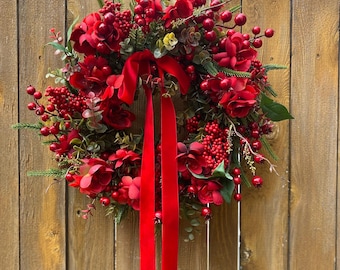 Könnte beinhalten: Ein runder Kranz mit dunkelroten Blumen, Büscheln aus kleinen roten Beeren und grünen Tannennadeln. Ein langes, leuchtend rotes Samtband ist oben in der Mitte gebunden, wobei zwei lange Bänder aus der Mitte des Kranzes herabhängen.